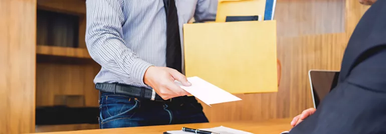 An employee, wearing a shirt and tie, extends a letter of resignation to their boss while holding a box of documents.